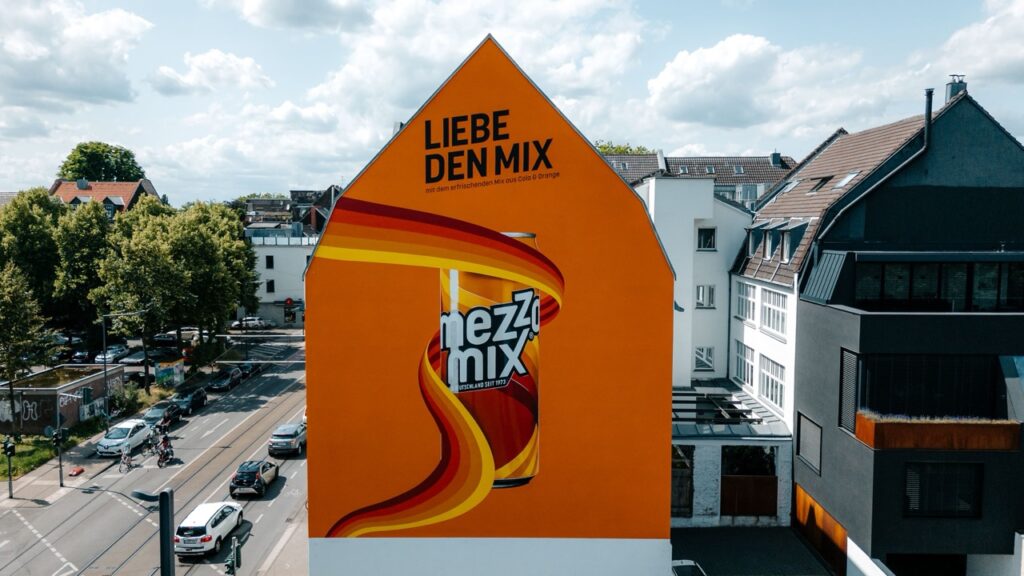 Großformatiges Wandbild mit Mezzo-Mix-Dose und Schriftzug „Liebe den Mix“ an einer Häuserwand in Köln-Ehrenfeld, handgemalt von Concrete Candy als Teil des Coca-Cola Changing Mural Projekts.