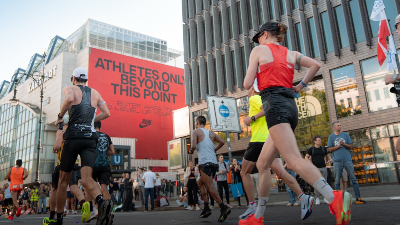 Läuferinnen und Läufer passieren während eines Marathons eine urbane Straßenszene in Berlin, im Hintergrund ein großflächiges rotes Nike-Takeover mit der Aufschrift „Athletes Only Beyond This Point“.
