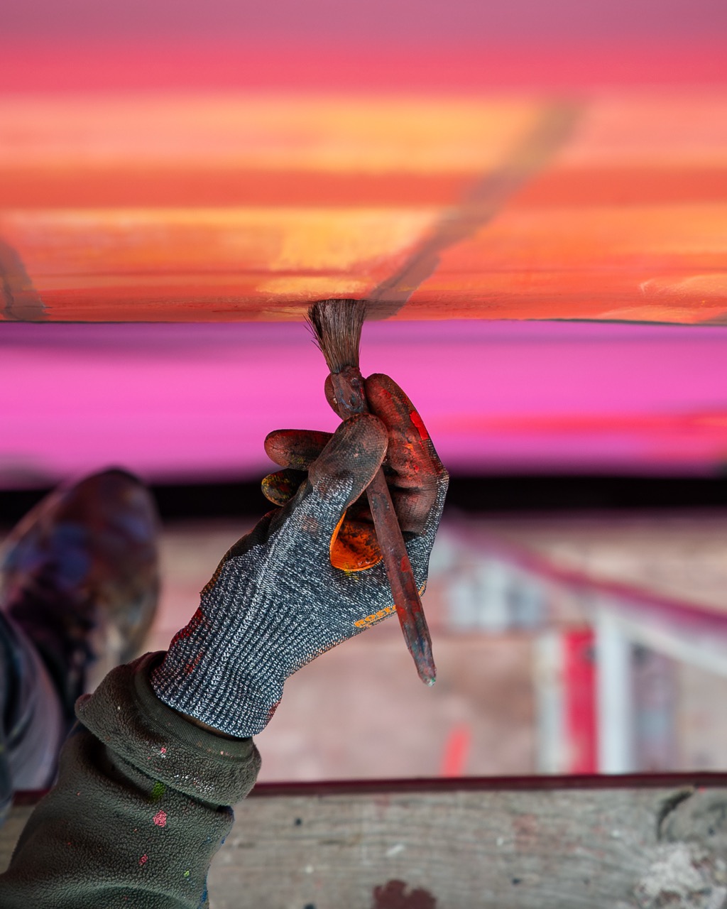 Detailaufnahme eines Künstlers beim Streichen mit Pinsel unter einer Konstruktion mit pink-orangefarbenem Farbverlauf