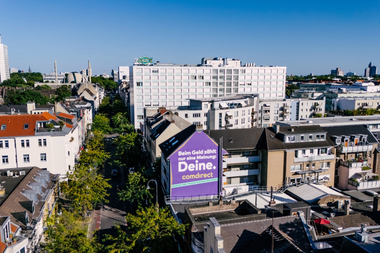 Seitliche Drohnenaufnahme eines violetten Comdirect Murals auf einer Giebelwand in Köln