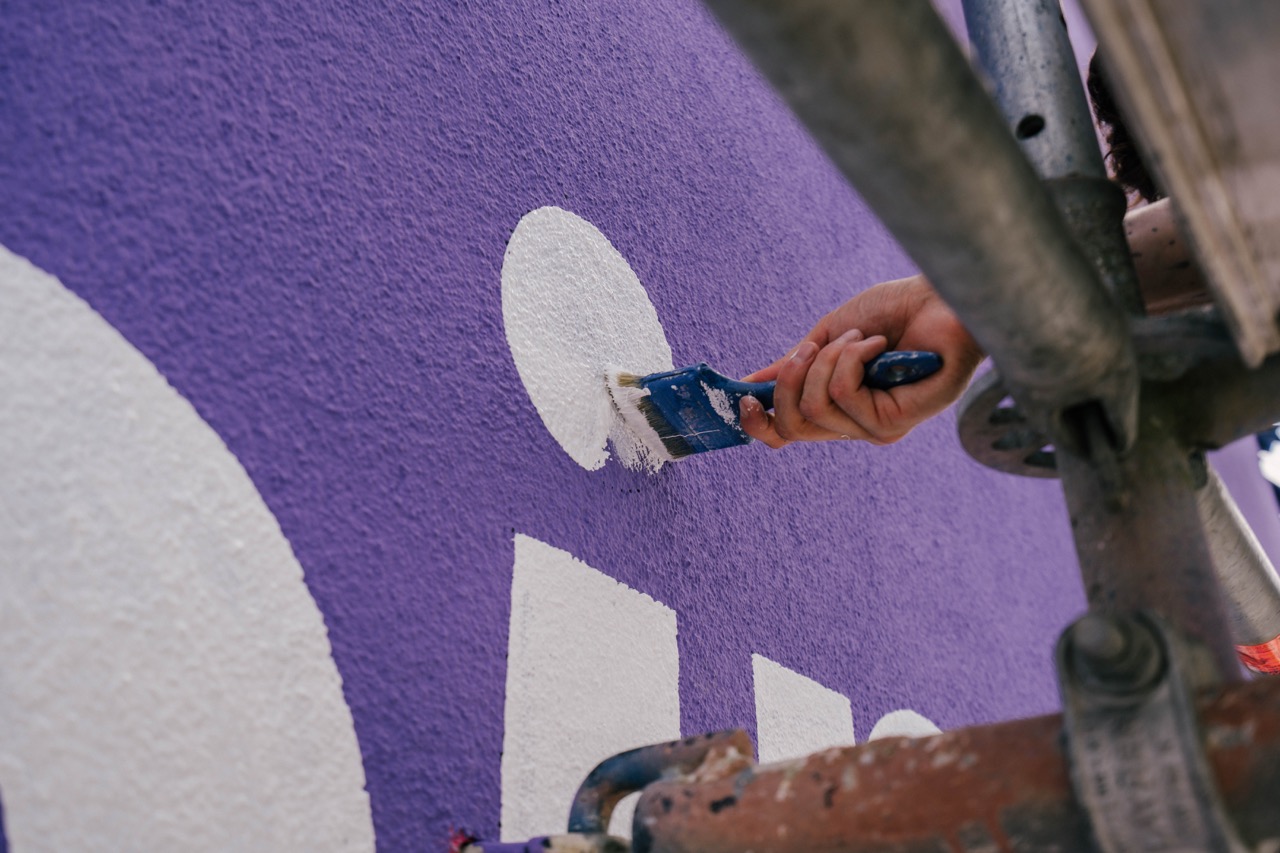 Detailaufnahme eines Pinsels beim Ausmalen weißer Buchstaben auf violettem Hintergrund