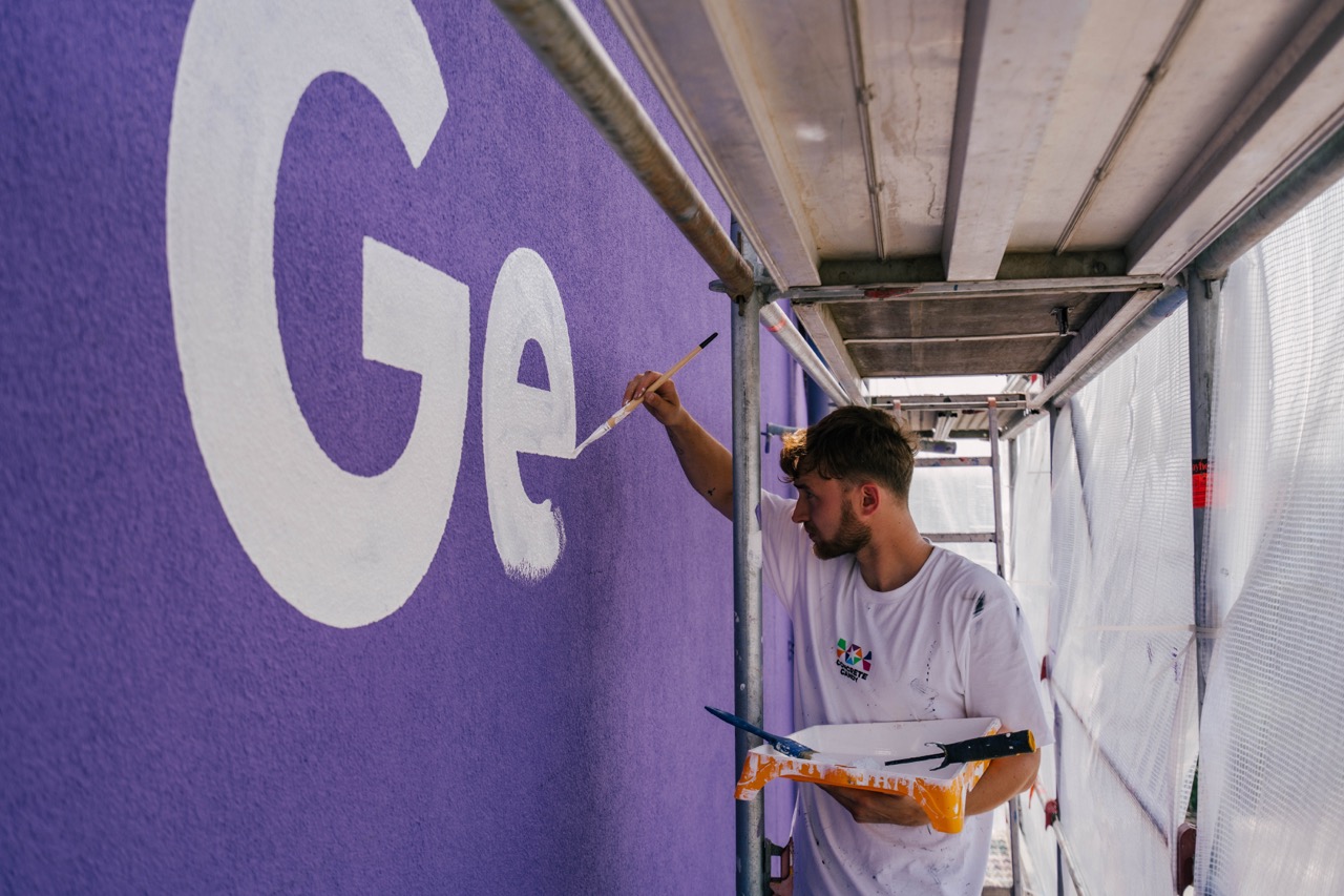 Künstler malt Buchstaben per Hand auf violette Hausfassade auf einem Gerüst
