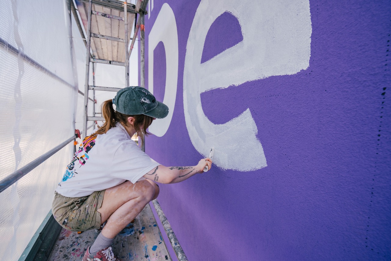 Künstlerin arbeitet an großformatigen weißen Buchstaben auf violettem Mural
