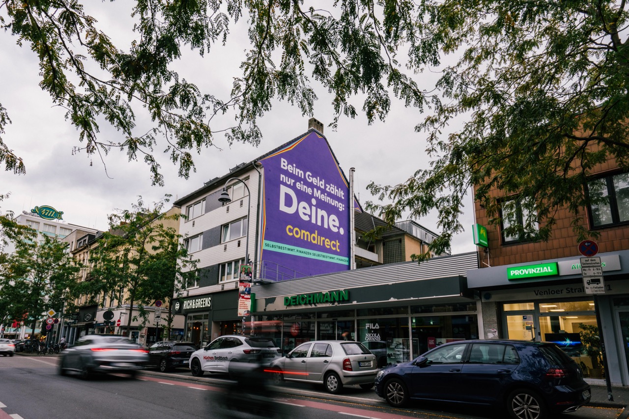 Straßenansicht des Comdirect Murals an der Venloer Straße in Köln mit vorbeifahrenden Fahrrädern und Autos