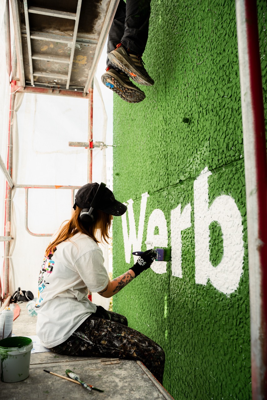 Künstlerin auf einem Gerüst beim handgemalten Lettering eines großformatigen S-Bahn München Murals an einer stark befahrenen Straße in München.