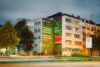 Nachtaufnahme des beleuchteten S-Bahn München Murals mit Lichtspuren vorbeifahrender Autos und urbaner Wohnbebauung in München.