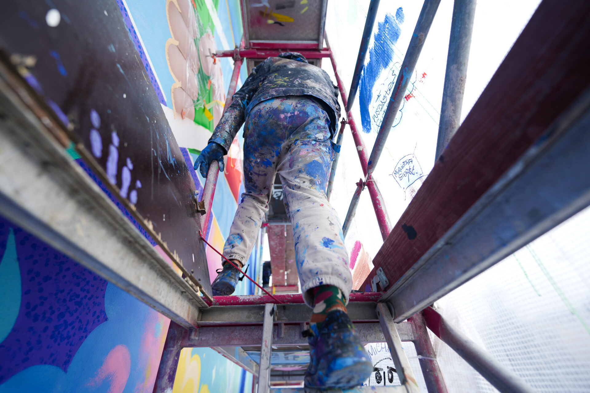 Künstler auf Gerüst arbeitet an großflächigem Firefox Mural, Low-Angle-Perspektive mit farbverschmierter Kleidung und dynamischen Formen an der Wand.