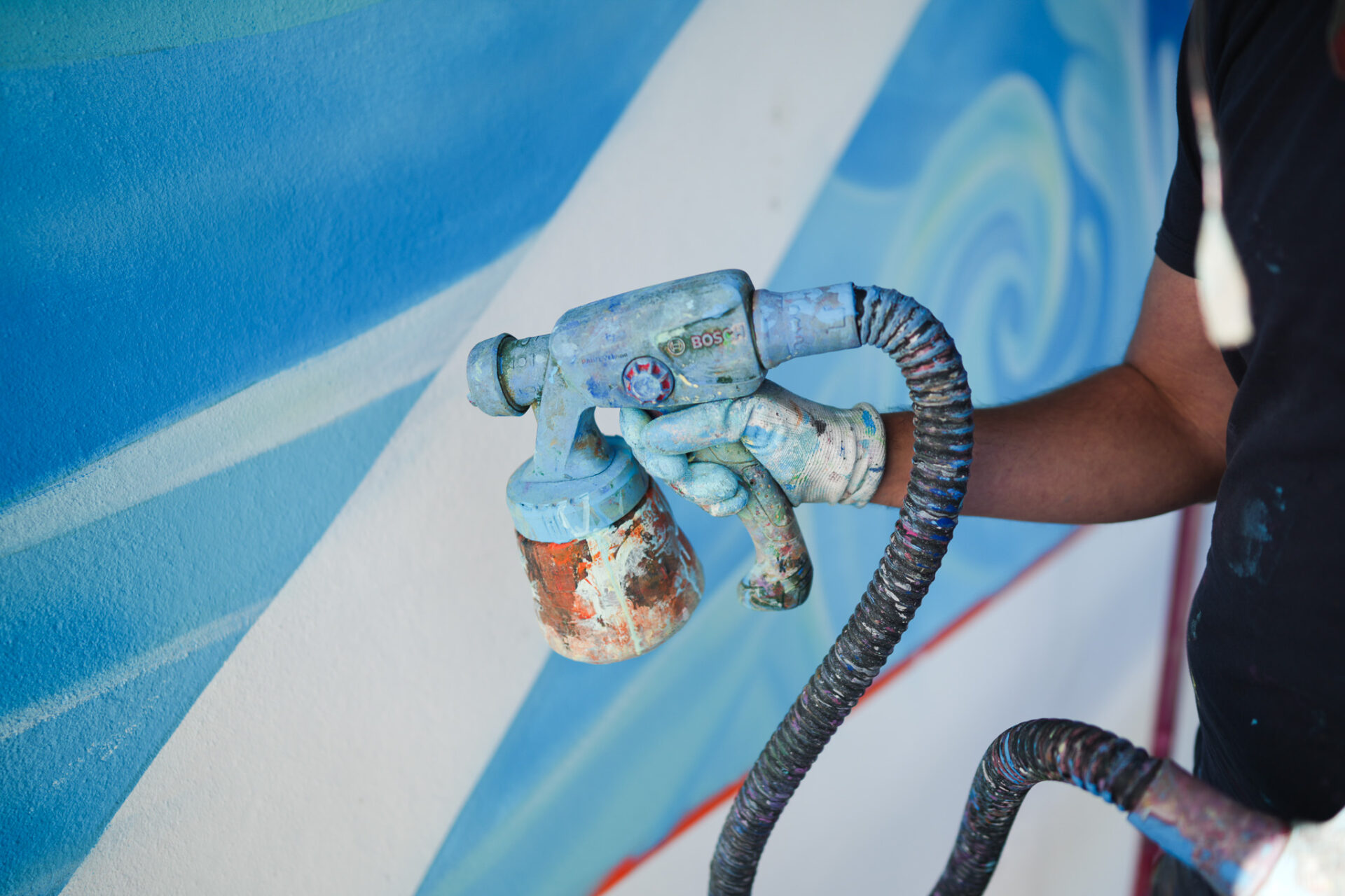 Detailaufnahme einer farbverschmierten Sprühpistole beim Auftragen weicher Farbverläufe auf eine Wand im Rahmen eines großformatigen Murals.