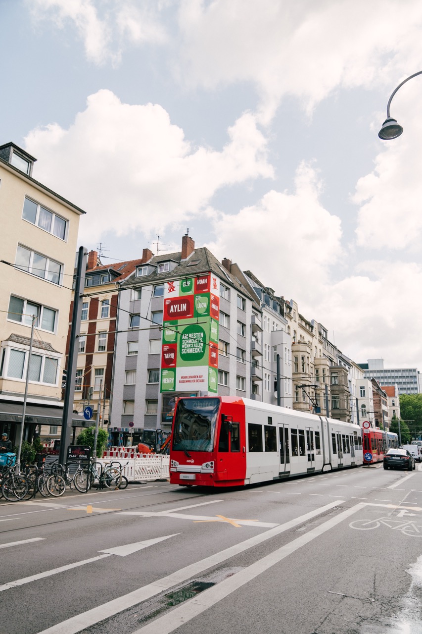 Straßenszene in Köln mit Straßenbahn im Vordergrund und großem Mural an einer Hauswand im Hintergrund.