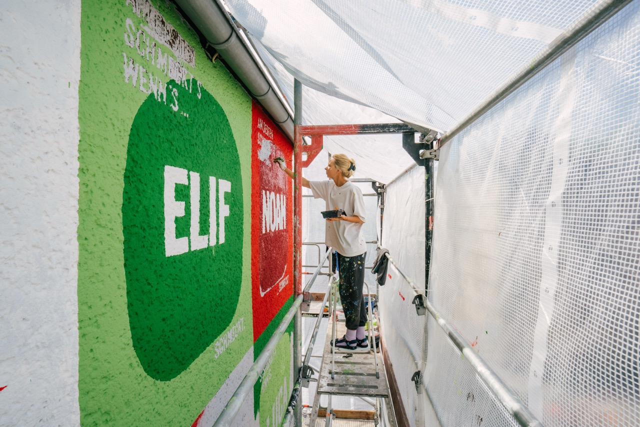Künstlerin malt auf einem Gerüst farbige Flächen und Schriftzüge an einer Hausfassade.