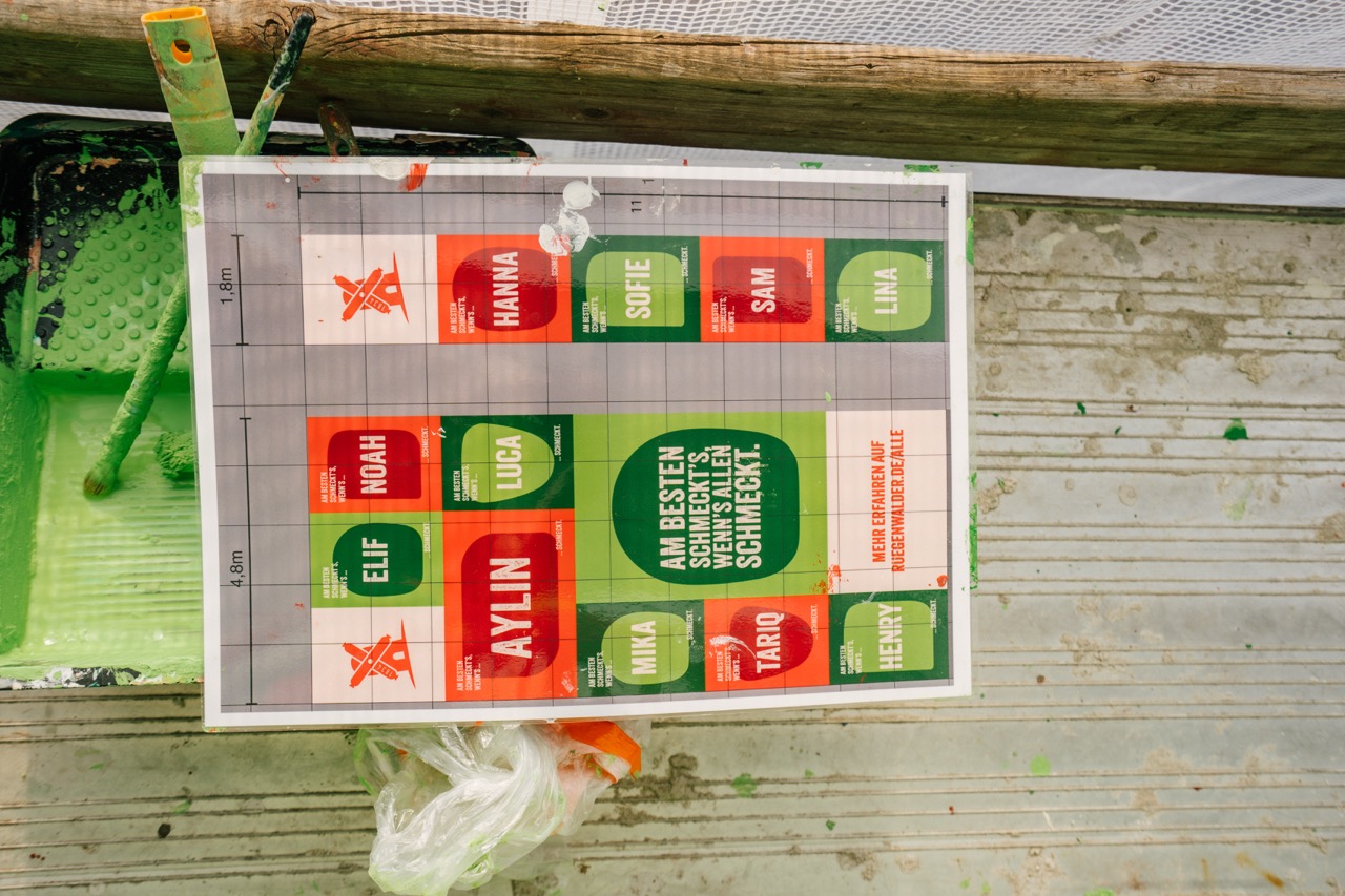 Ausgedruckter Entwurf eines Murals mit farbigen Flächen und Namen liegt auf einem Gerüst.