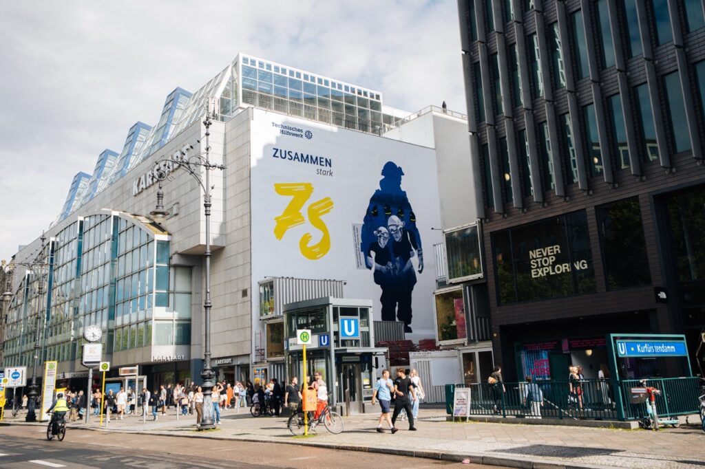 Großes THW-Mural an einer Gebäudefassade am Kurfürstendamm mit vorbeilaufenden Menschen im Vordergrund.