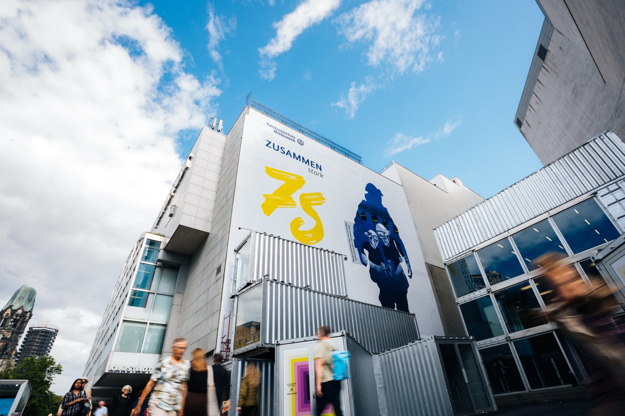 Untersicht eines THW-Murals an einer hohen Gebäudefassade mit vorbeiziehenden Menschen im Vordergrund.