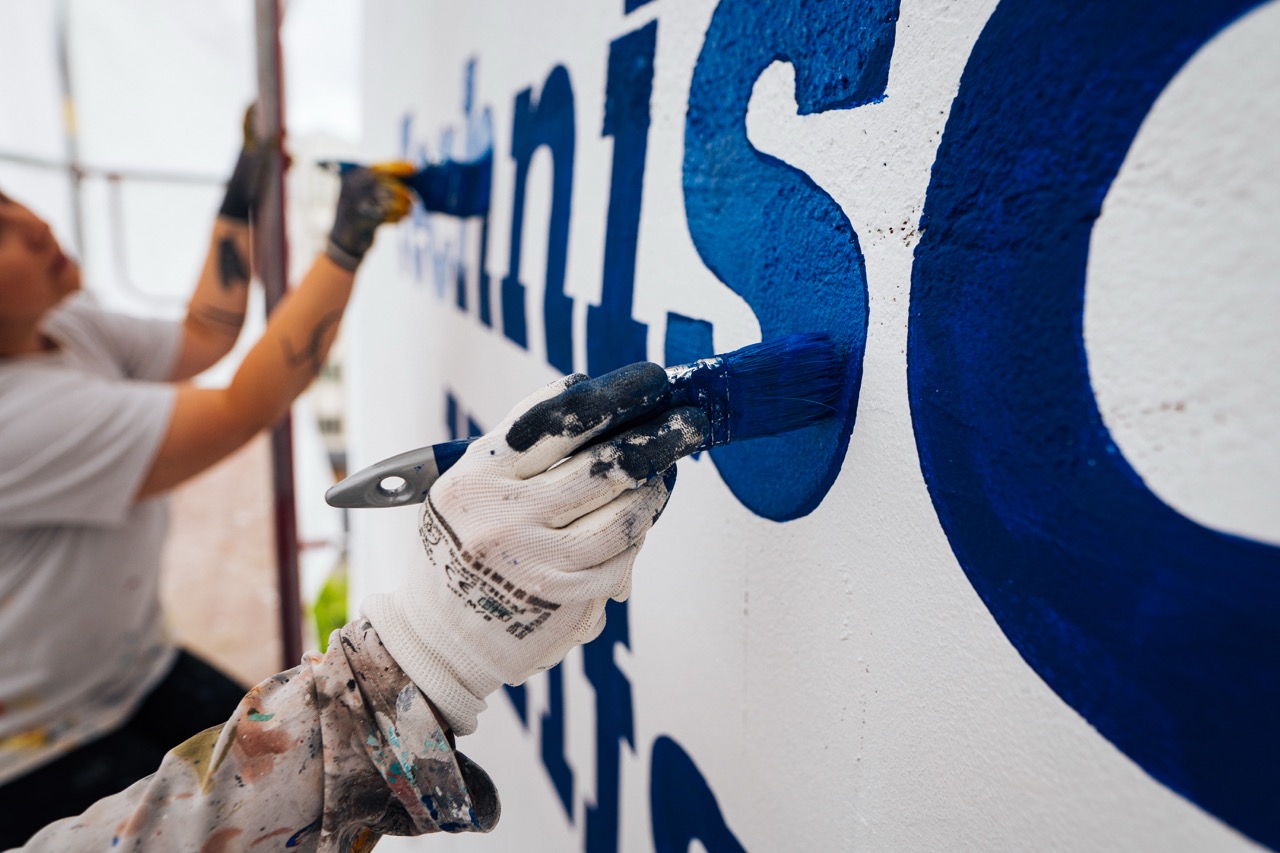 Detailaufnahme eines Handschuhs, der blaue Schriftzüge präzise mit dem Pinsel auf eine weiße Wand malt.