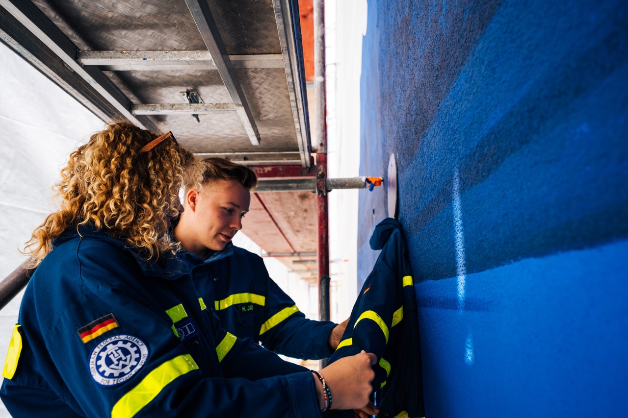 Zwei Personen in THW-Uniform befestigen Kleidung an einer blau gestrichenen Wand auf einem Gerüst.