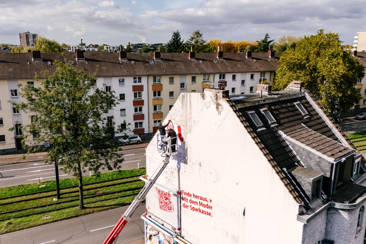 Hebebühne mit zwei Personen beim Anbringen eines Murals an einer Giebelwand in einem Wohngebiet.