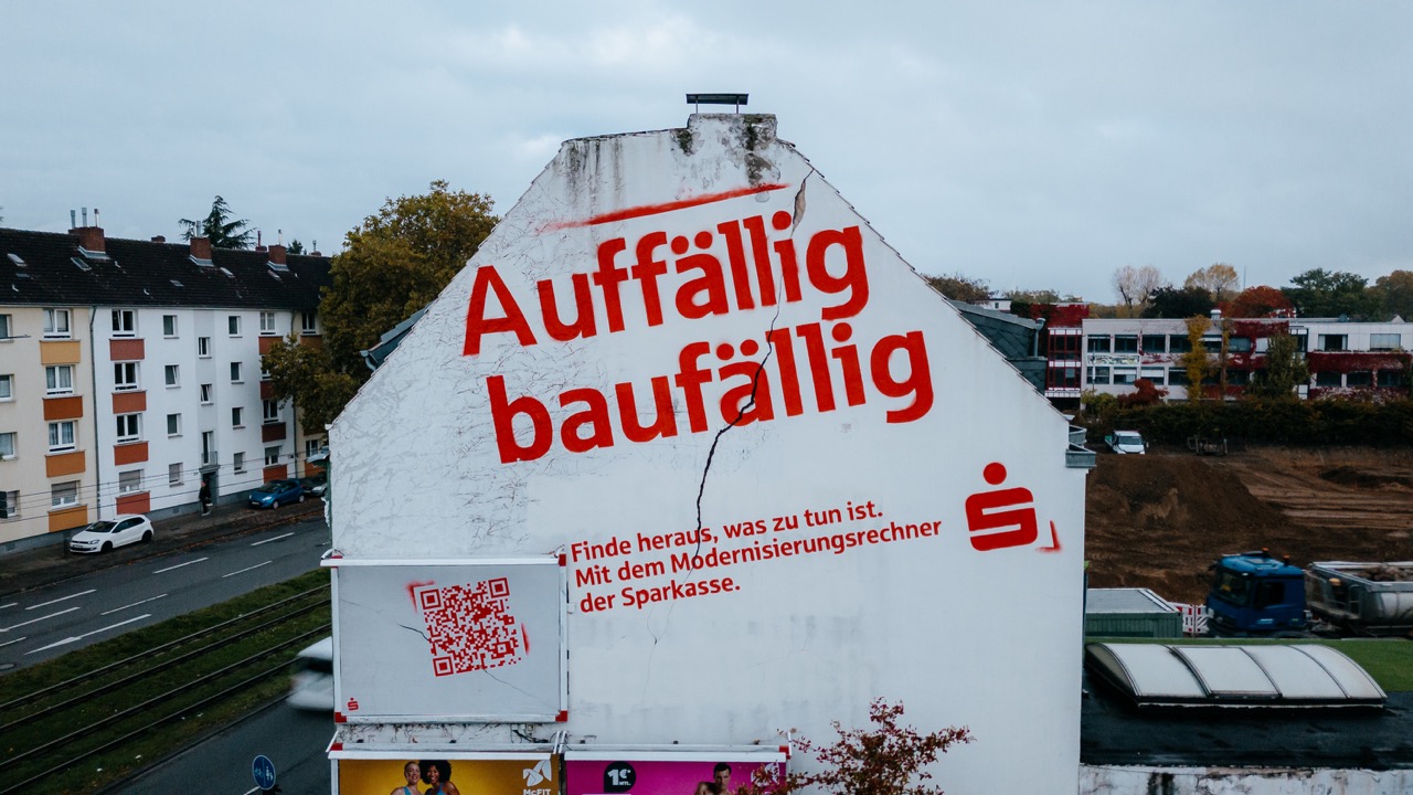 Großflächiges Sparkassen-Mural mit roter Schrift „Auffällig baufällig“ auf einer Hausfassade.