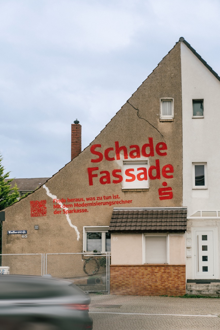 Hausfassade mit rotem Schriftzug „Schade Fassade“ und QR-Code als Teil einer Sparkassen-Kampagne.