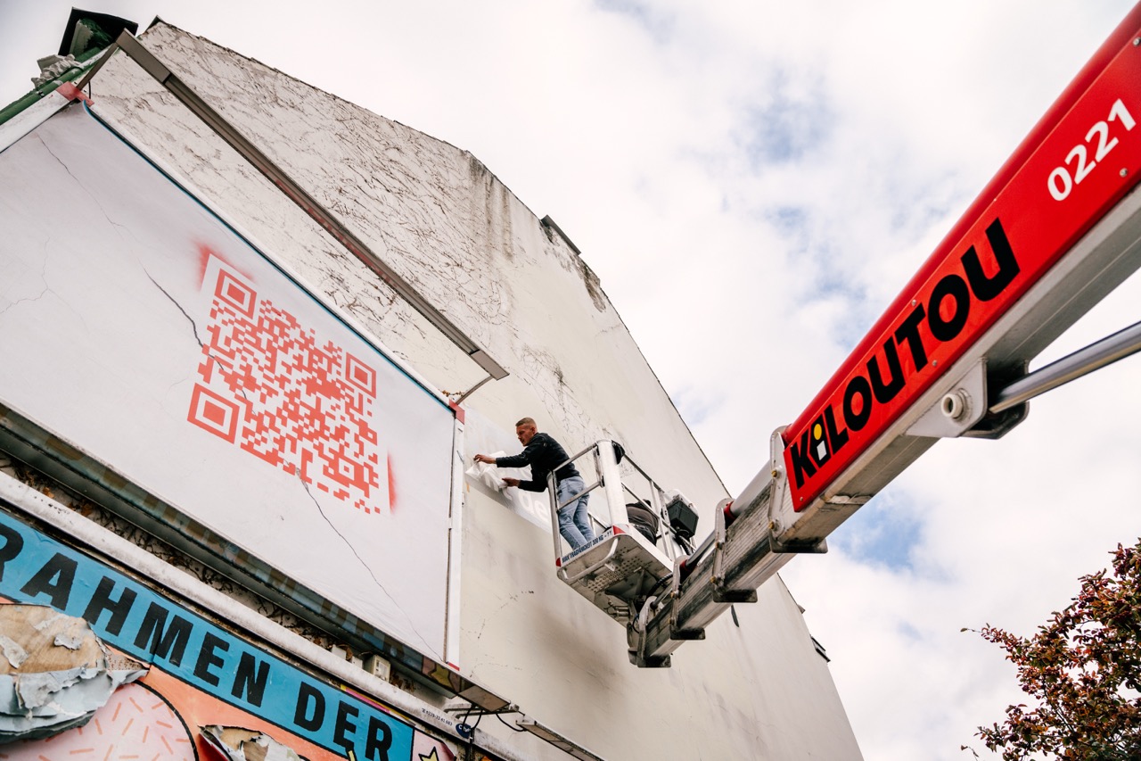 Person arbeitet auf einer Hebebühne an einer Hauswand neben einem großen QR-Code und Sparkassen-Branding.