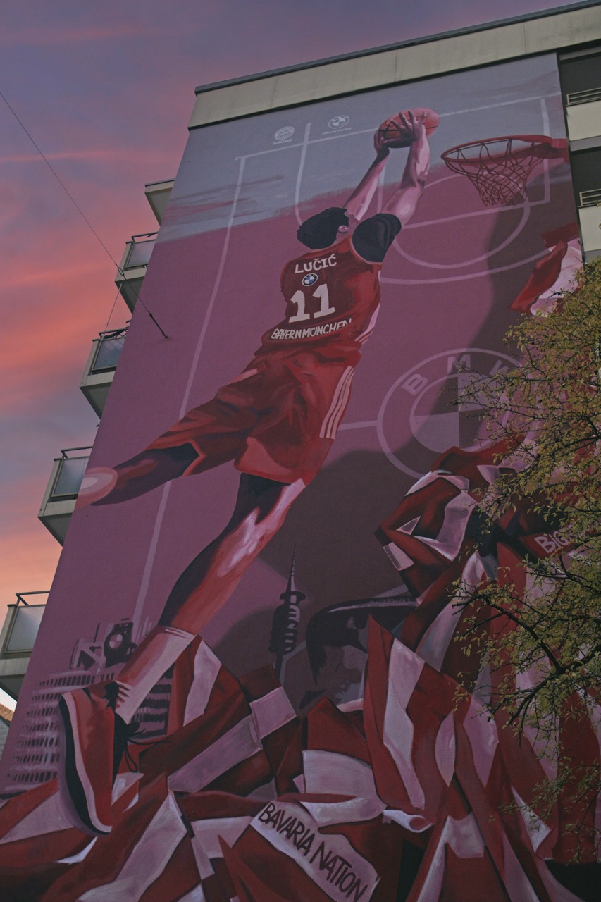 Detailaufnahme des FC Bayern Basketball Murals mit springendem Spieler, Basketballkorb und rot-weißen Fanflaggen bei Abendstimmung.