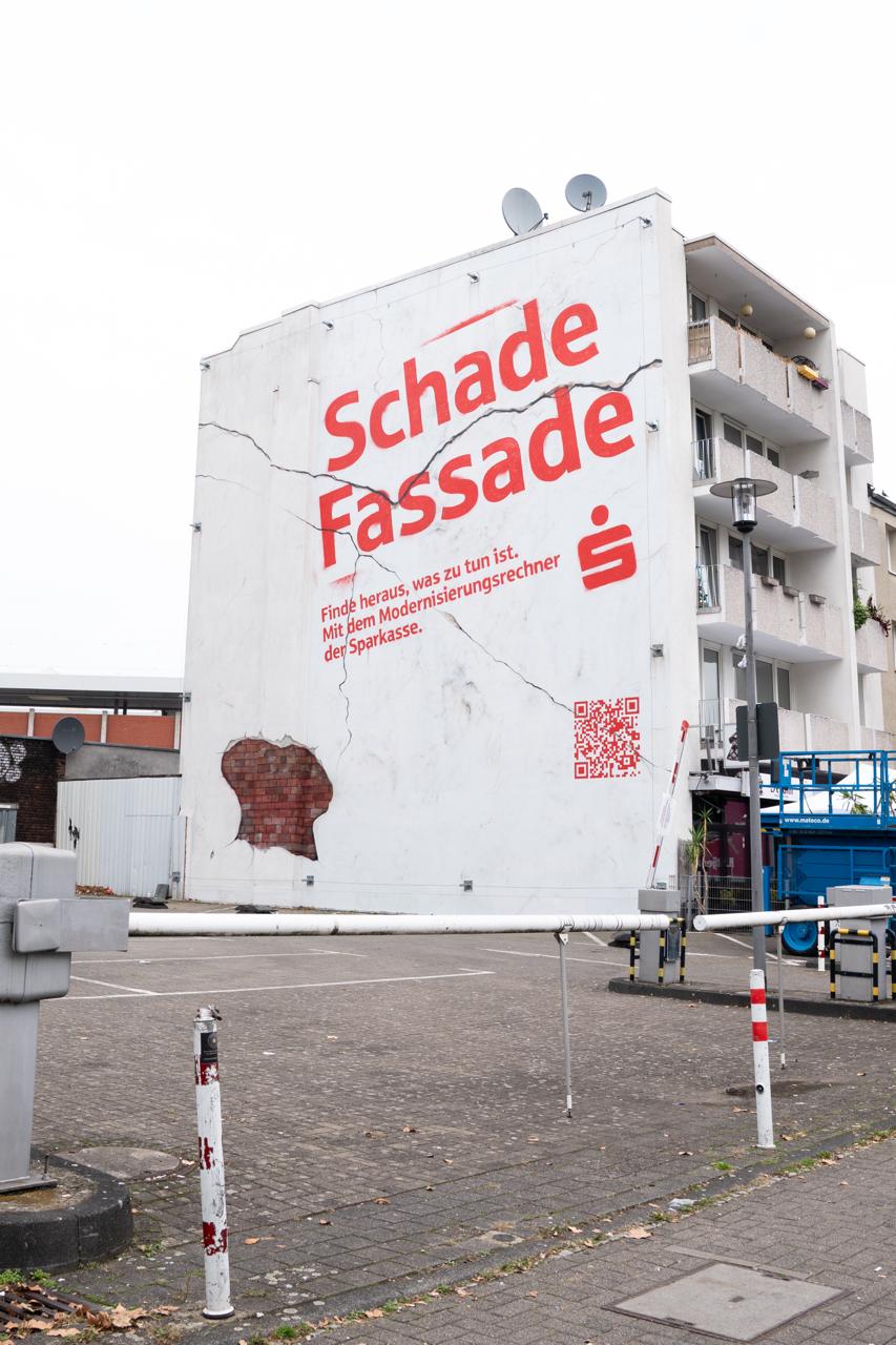 Großes Sparkassen-Mural mit Schriftzug „Schade Fassade“ und sichtbaren Rissen in der Gebäudewand.