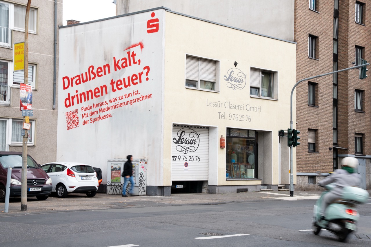 Sparkassen-Mural mit Text „Draußen kalt, drinnen teuer?“ an einer Gebäudeecke im urbanen Umfeld.