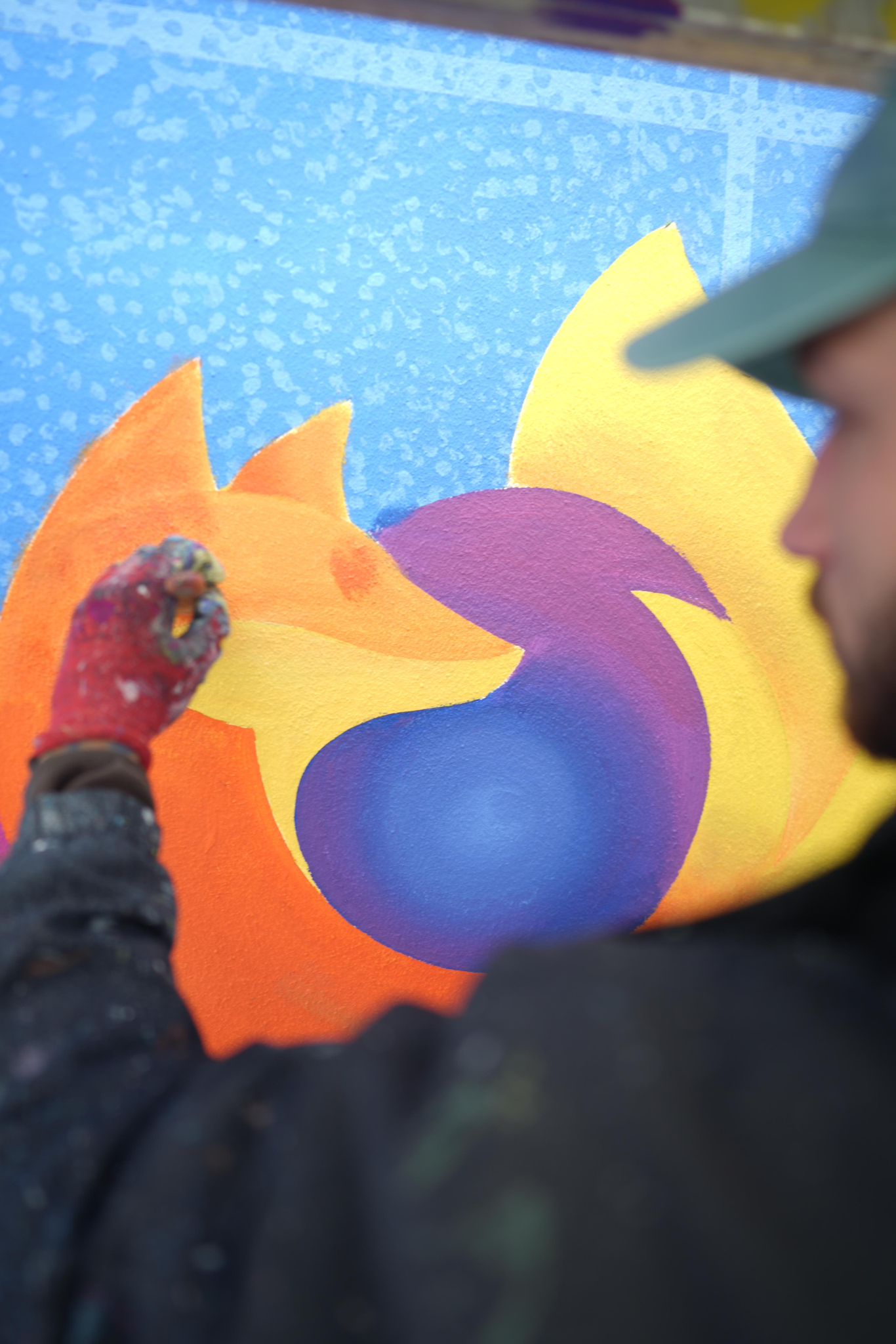 Künstler arbeitet an leuchtenden Farbverläufen in Gelb, Orange und Violett auf einer strukturierten Wandoberfläche.