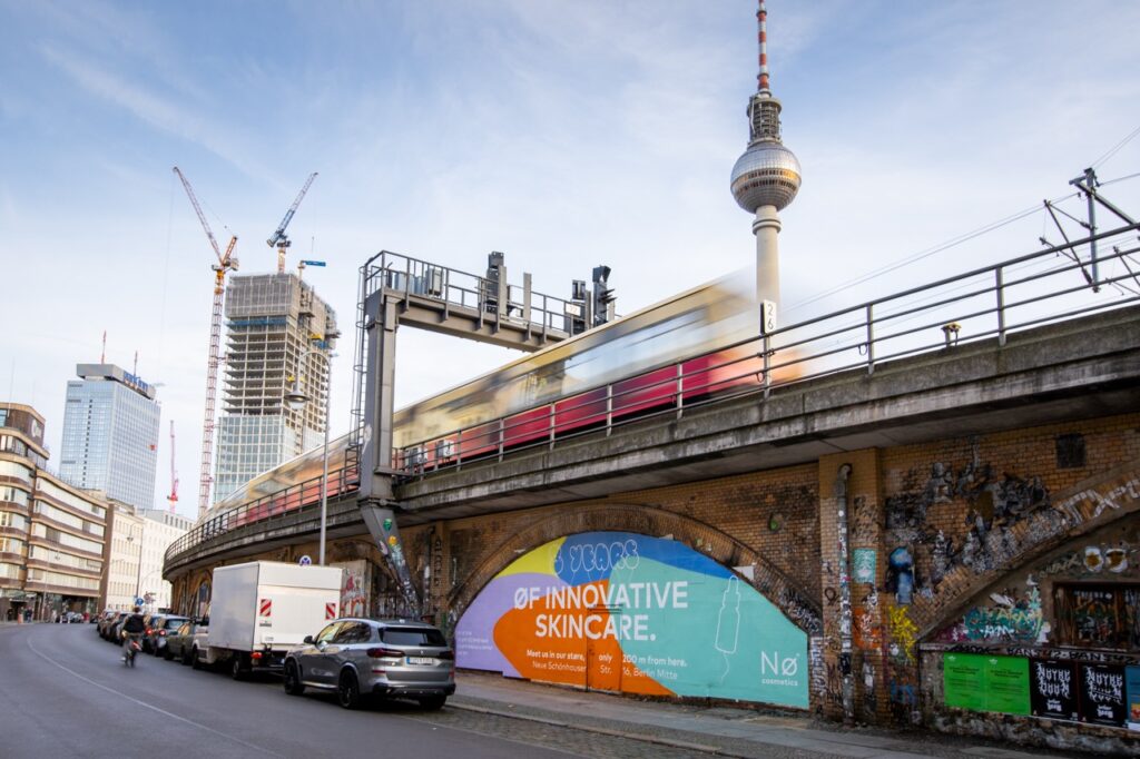 Nø Cosmetics Kampagne an der Dircksenstraße in Berlin mit vorbeifahrender S-Bahn, Fernsehturm und urbaner Architektur im Hintergrund.