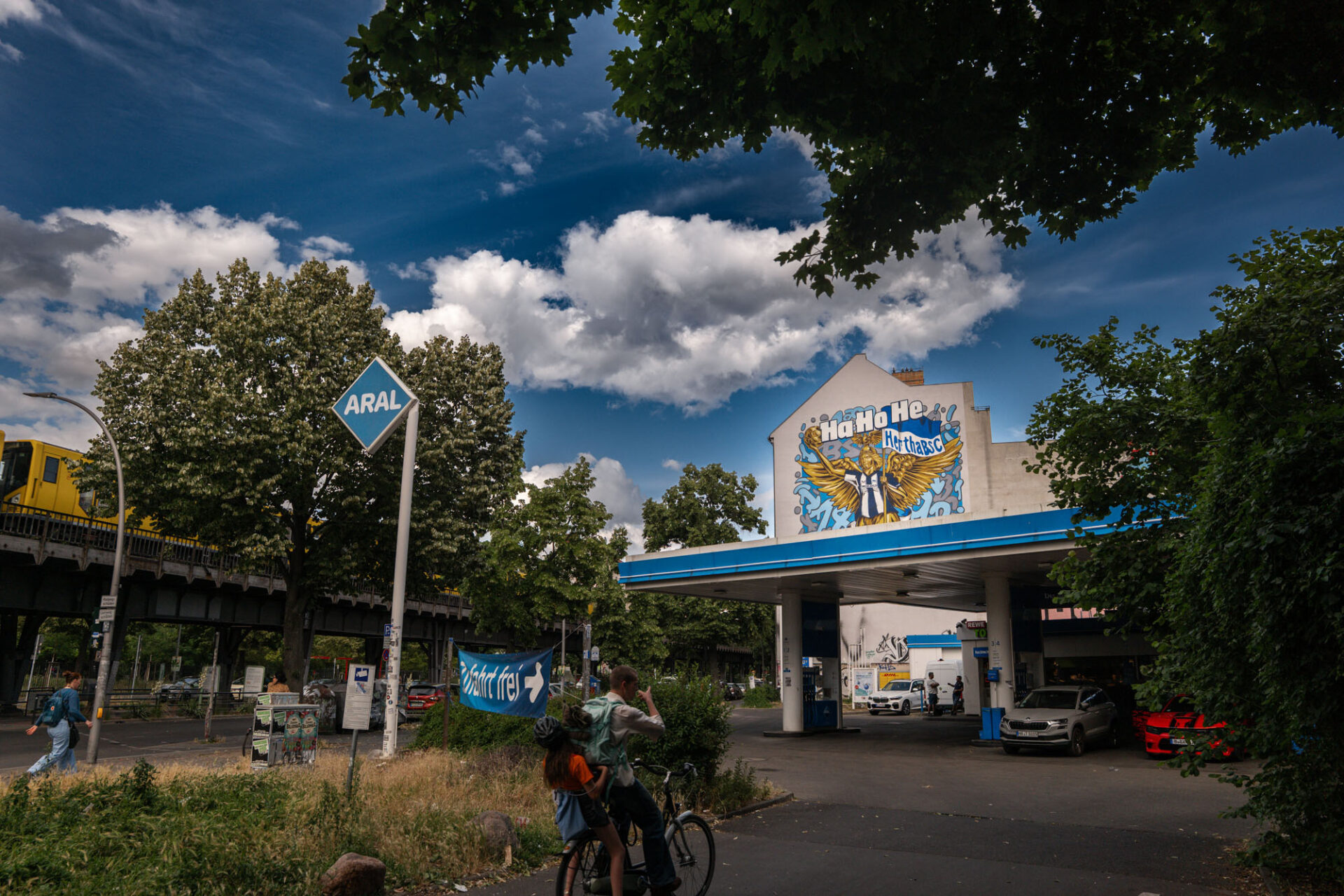 Weitwinkelaufnahme eines Hertha-BSC-Murals an einer Gebäudefassade neben Bahntrasse und Tankstelle in Berlin.