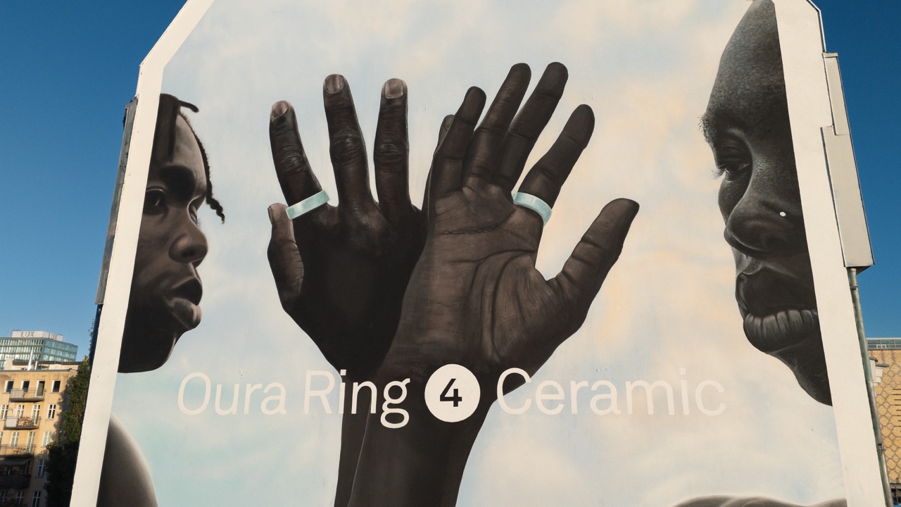 Großflächiges Mural am East Side Hotel in Berlin mit zwei realistisch dargestellten Gesichtern und erhobenen Händen, die den Oura Ring 4 Ceramic tragen; zentral platziert vor hellem Hintergrund mit Markeninschrift.