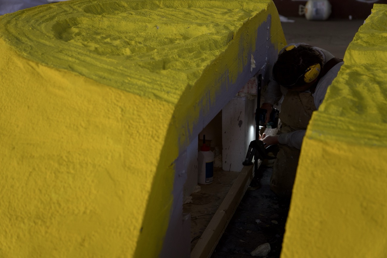 Person arbeitet in einer Werkhalle an einem großen gelben 3D-Objekt und bearbeitet es mit einem Werkzeug, während Schutzkleidung und Gehörschutz getragen werden.