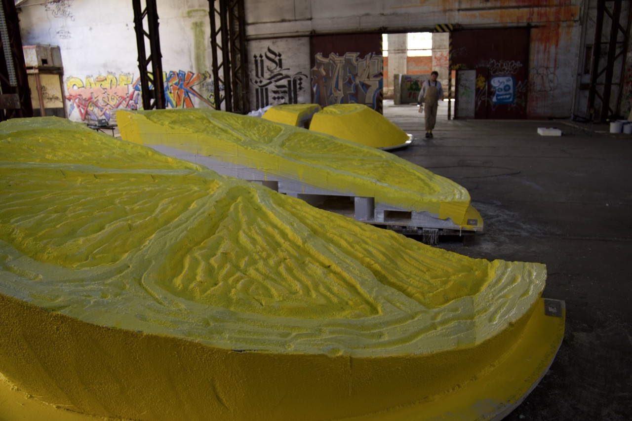 Mehrere große, gelb gestrichene 3D-Zitronenformen liegen in einer industriellen Halle mit Graffiti im Hintergrund und werden für den Einsatz im Mural vorbereitet.