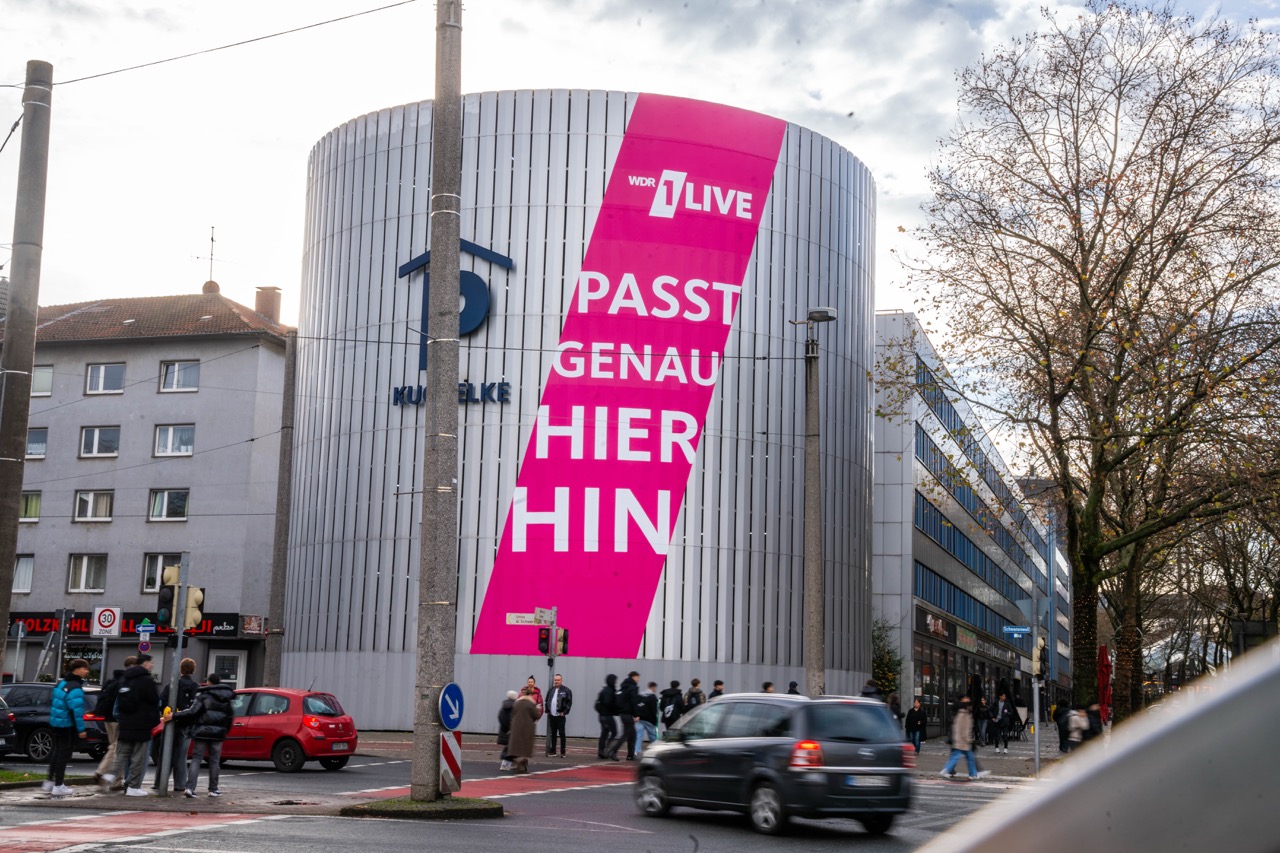 Großformatige 1LIVE Kampagne mit pinkem Banner und weißer Typografie an einem Parkhaus in Dortmund aus Straßenperspektive
