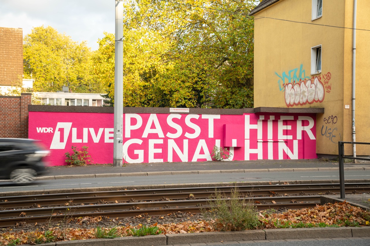 Seitliche Ansicht eines langen 1LIVE Murals in Gelsenkirchen entlang einer Straße mit vorbeifahrendem Auto