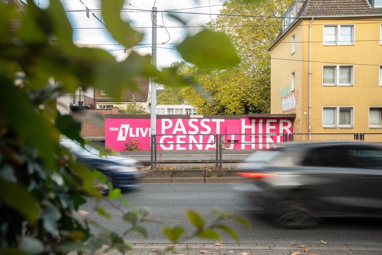 1LIVE Mural in Gelsenkirchen durch Blätter im Vordergrund und vorbeifahrende Autos eingerahmt