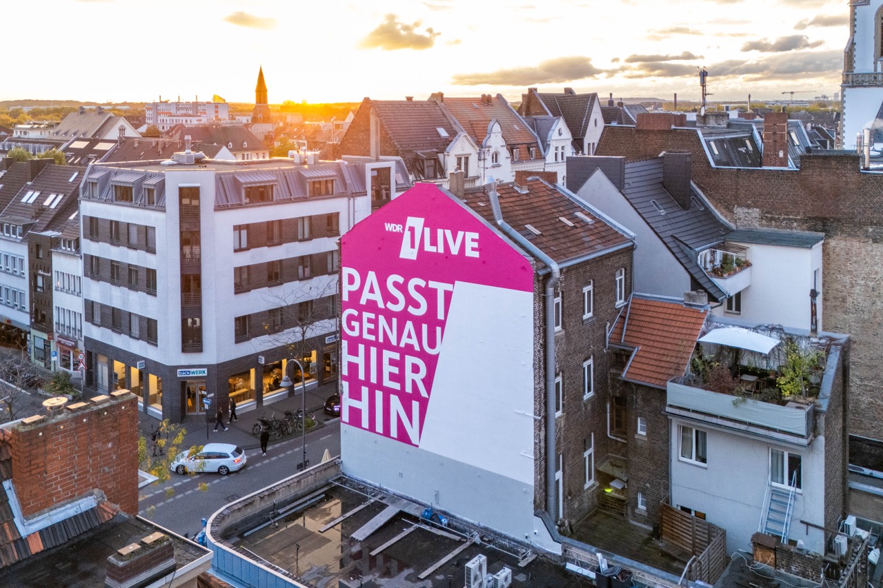 Großflächiges 1LIVE Mural an einer Hausfassade in Köln bei Sonnenuntergang mit pinker Typografie „Passt genau hier hin“