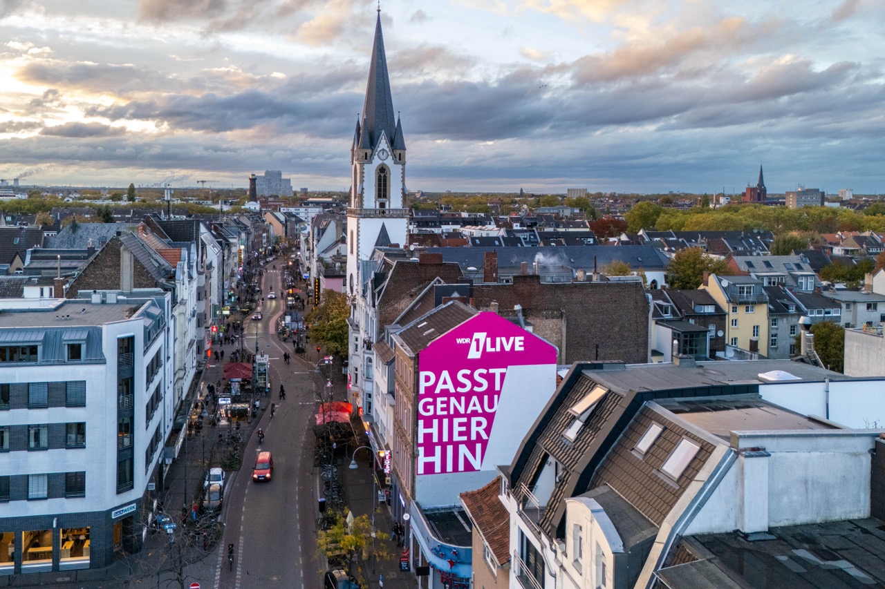 Weitwinkelaufnahme eines 1LIVE Murals in Köln mit Blick über die Stadt und Kirche im Hintergrund