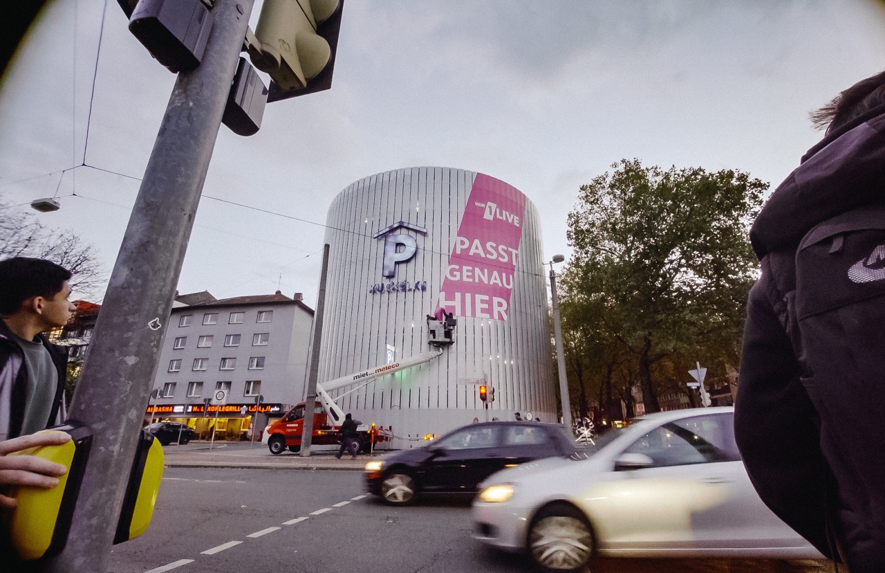 Montage einer großflächigen 1LIVE Beklebung an einem runden Parkhaus in Dortmund während des laufenden Straßenverkehrs