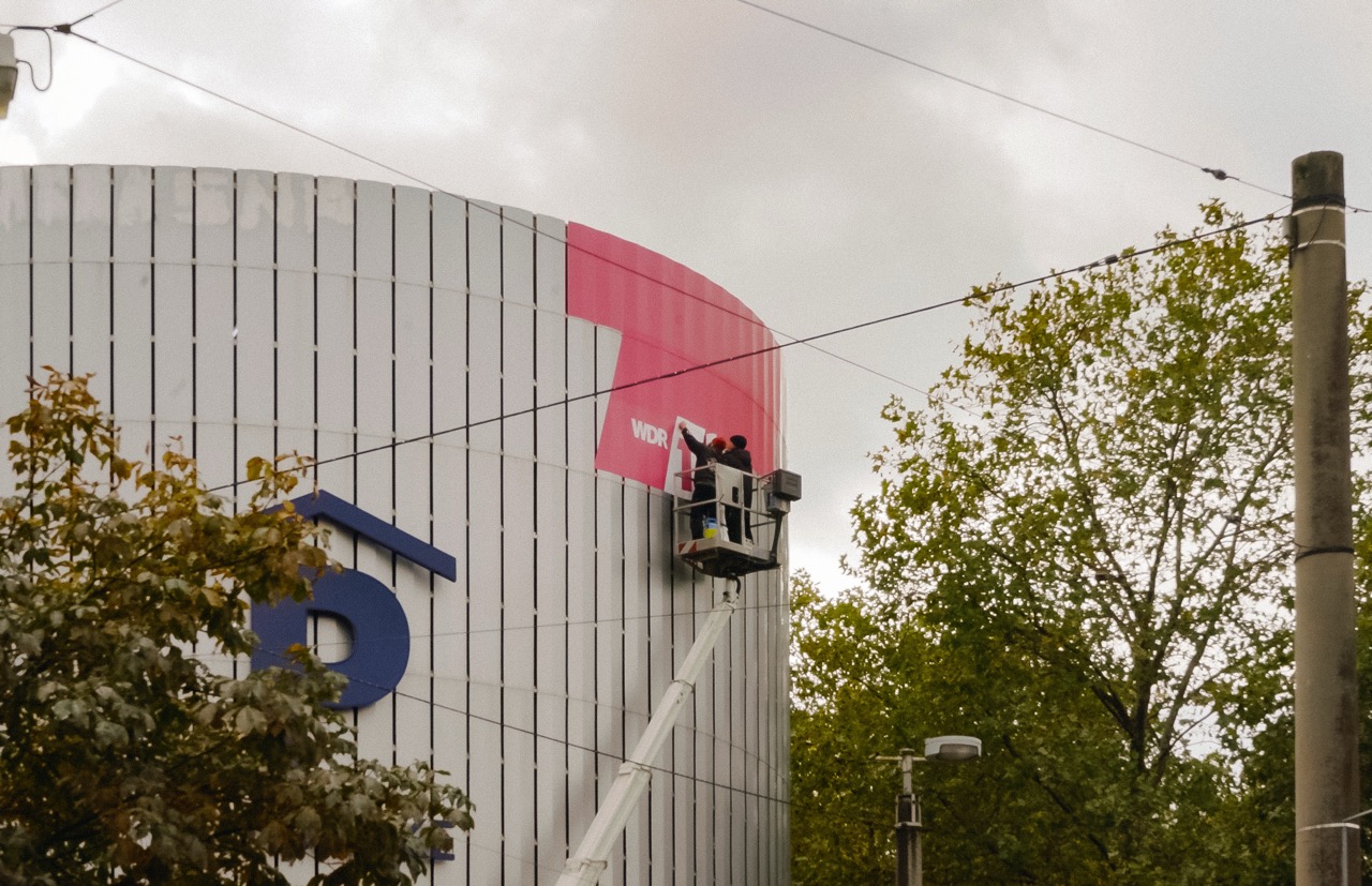 Zwei Arbeiter bringen eine pinke 1LIVE Folie mit Hebebühne an der Fassade eines Parkhauses in Dortmund an