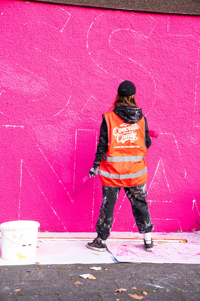 Künstlerin in Warnweste zeichnet die Buchstaben eines 1LIVE Murals auf eine pinke Wand