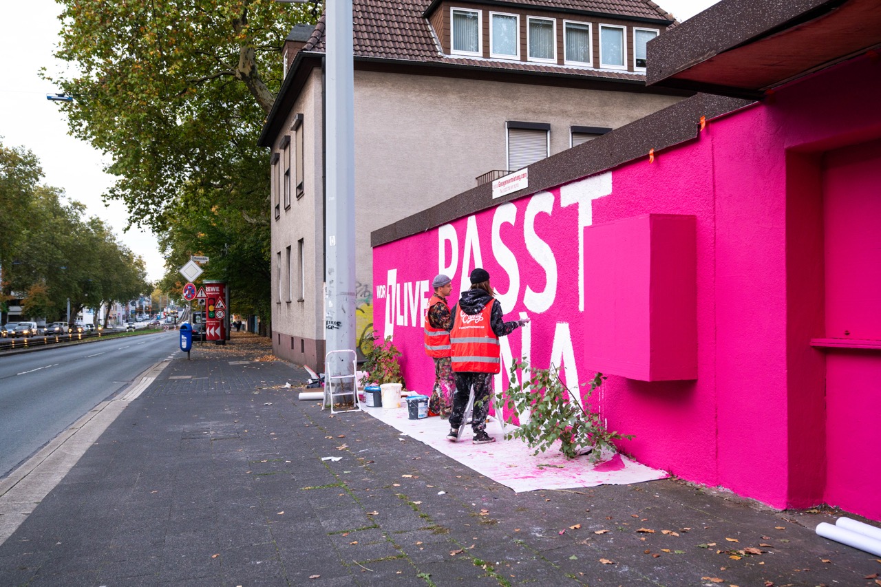Künstler arbeiten an den finalen Details eines pinken 1LIVE Murals entlang einer Straße in Gelsenkirchen