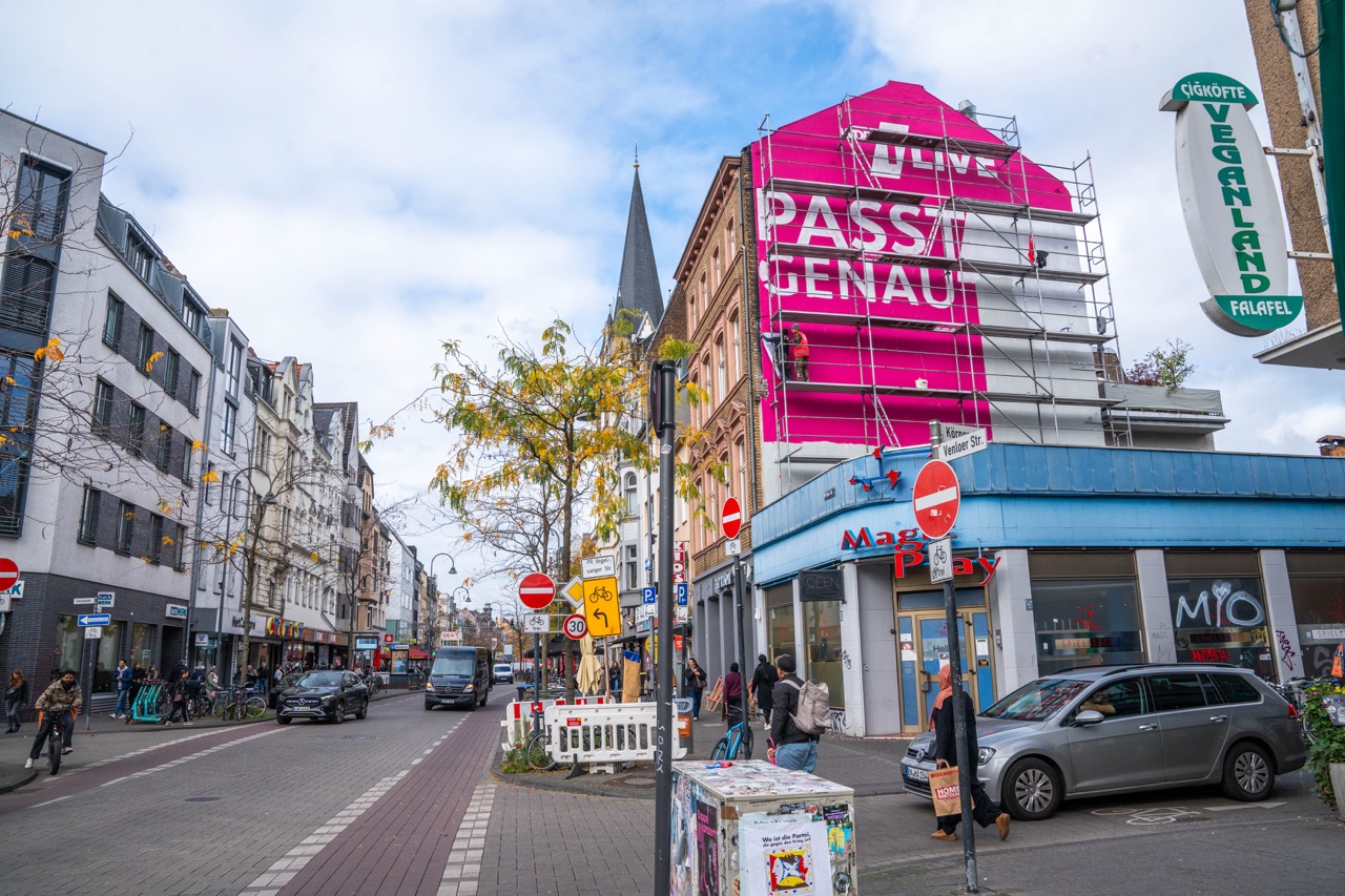 Großes 1LIVE Mural im Entstehungsprozess an einer Hausfassade in Köln mit Gerüst und Straßenleben im Vordergrund