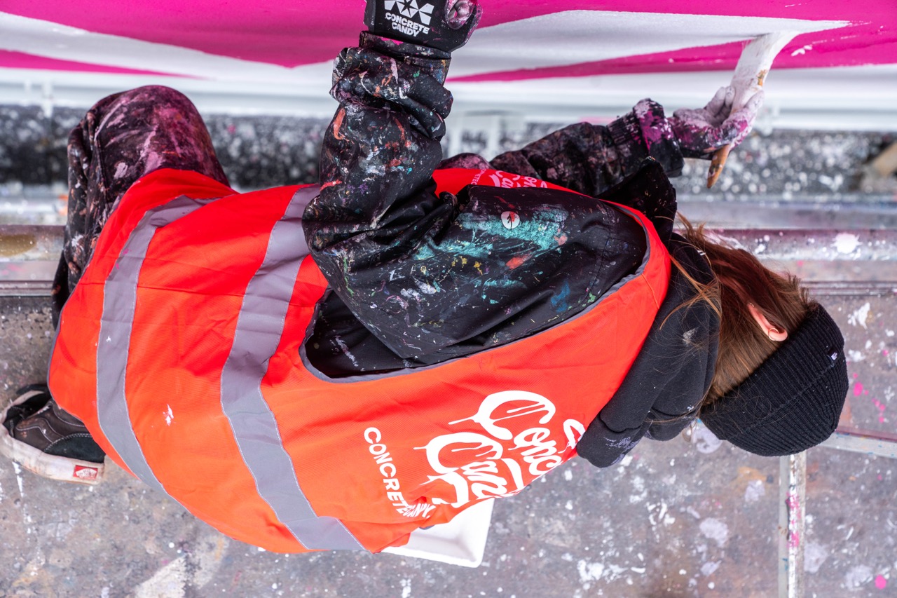 Künstlerin in Warnweste malt liegend auf einem Gerüst an einem pinken 1LIVE Mural