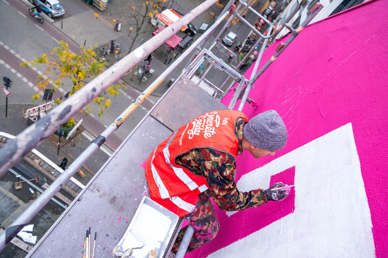 Künstler malt weiße Typografie auf pinkem Hintergrund eines 1LIVE Murals aus der Vogelperspektive