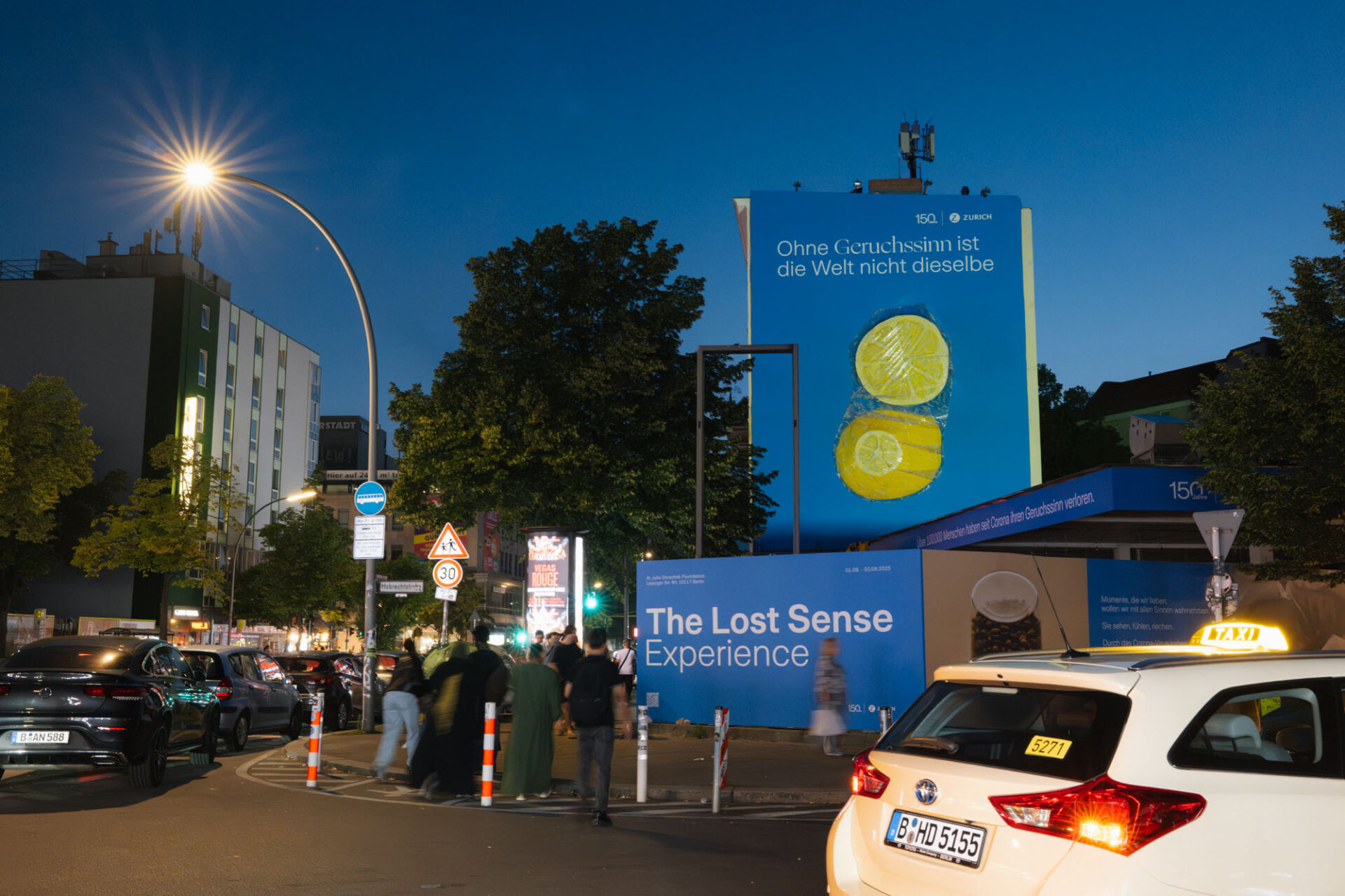 Belebte Straßenszene bei Nacht mit Menschen, Autos und einem großen Mural sowie einer Installation mit der Aufschrift „The Lost Sense Experience“.
