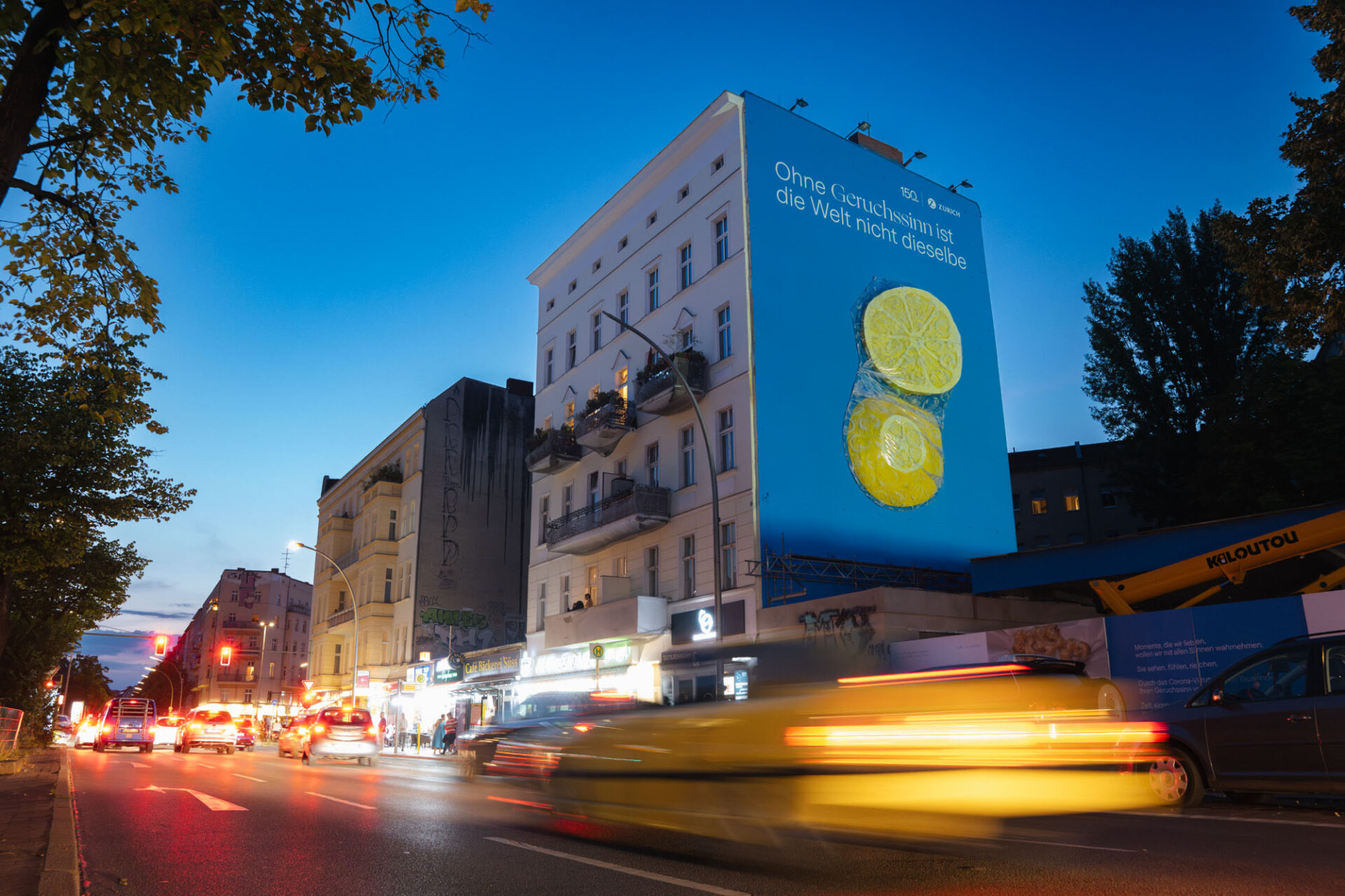 Nachtaufnahme einer Berliner Straße mit vorbeifahrenden Autos in Bewegung, während ein großformatiges Mural mit Zitronen auf einer Hauswand beleuchtet wird.