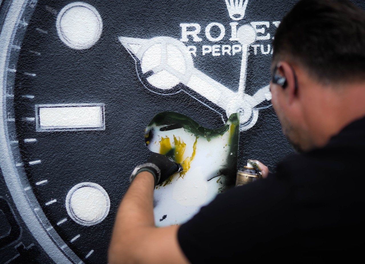 Künstler TANK arbeitet mit Schablone und Spraydose an feinen Details des Rolex Uhrmotivs auf der Wand.