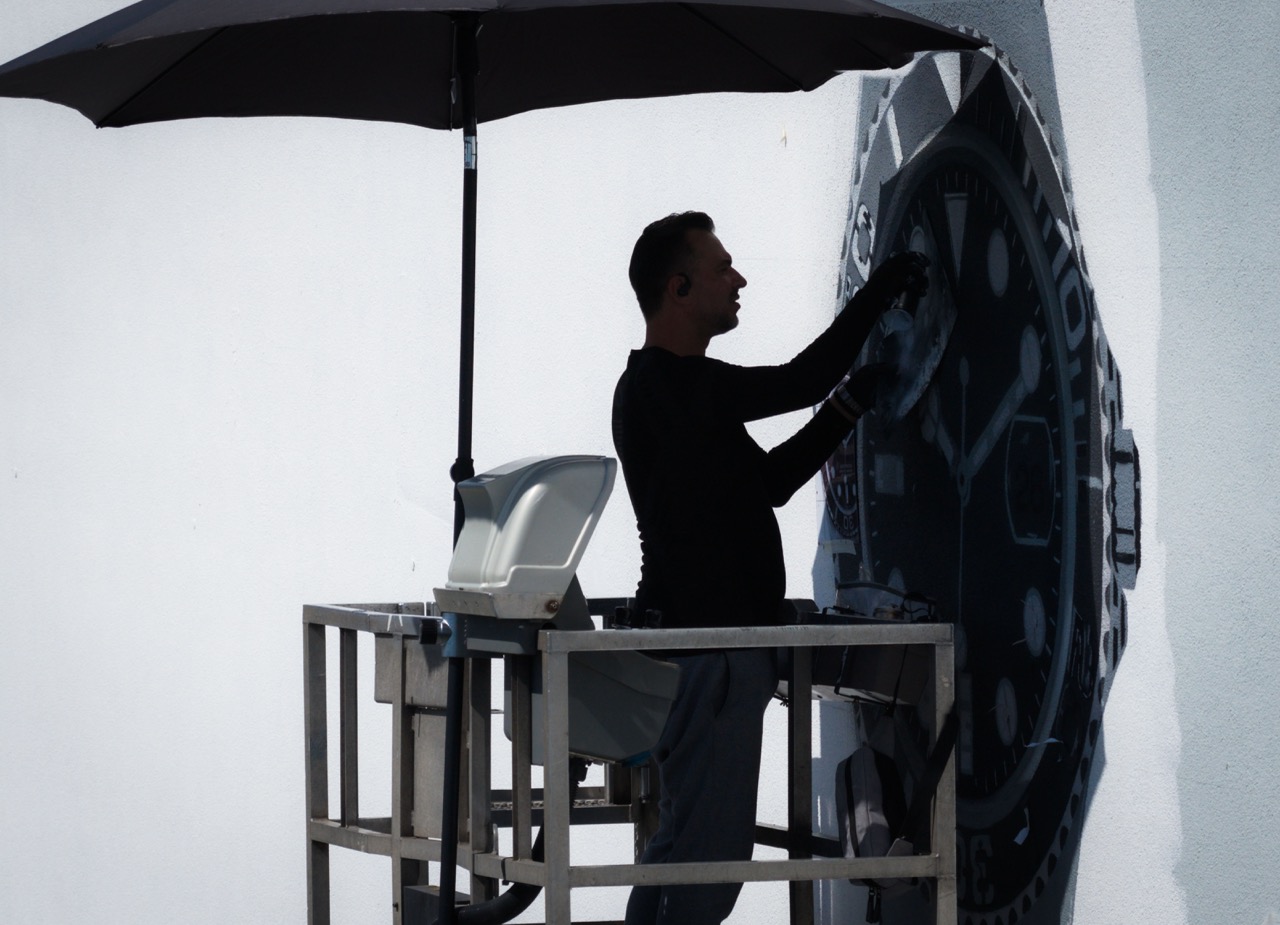 Künstler TANK malt in luftiger Höhe an einem überdimensionalen Rolex Zifferblatt, geschützt unter einem Sonnenschirm.