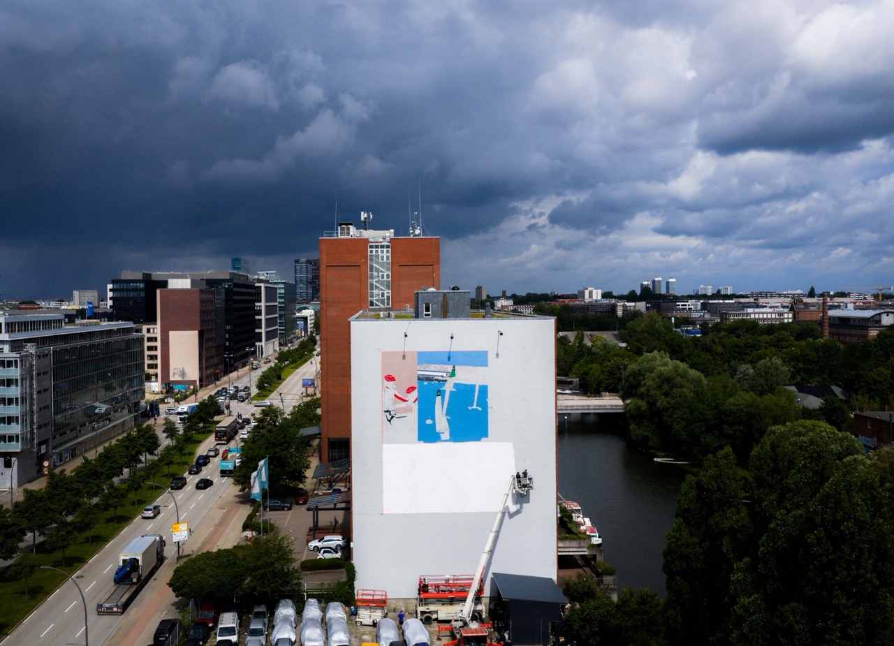 Großflächiges Mural im Aufbau an einer Gebäudefassade in Hamburg, mit Hebebühne und ersten farbigen Elementen des SailGP-Motivs.