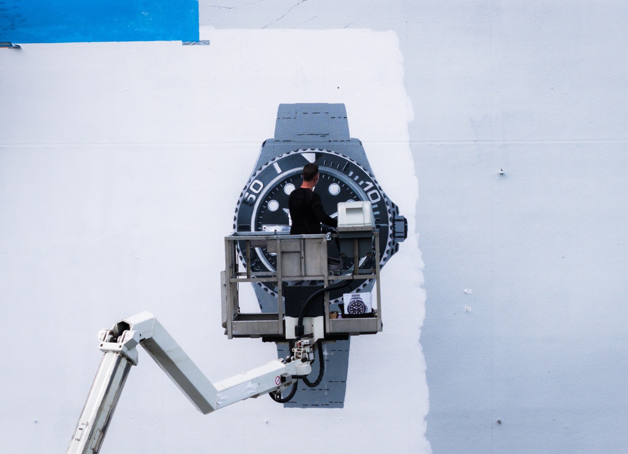 Zwei Künstler setzen letzte Details am fotorealistischen Rolex Mural mit feinen Linien und Highlights.